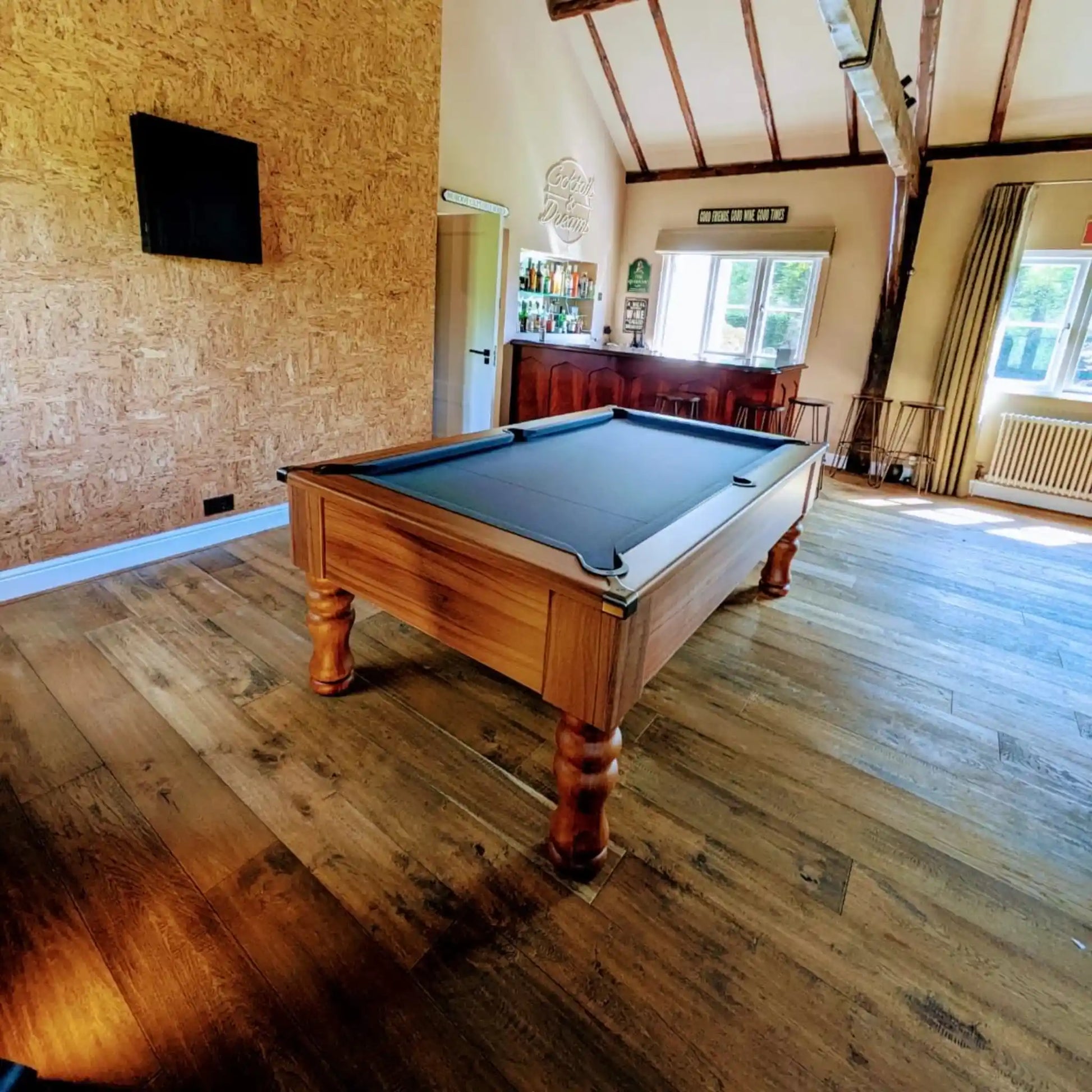 A wooden pool table with a dark green felt surface is shown in a large room with wood floors, exposed ceiling beams, windows, and a bar area in the background.
