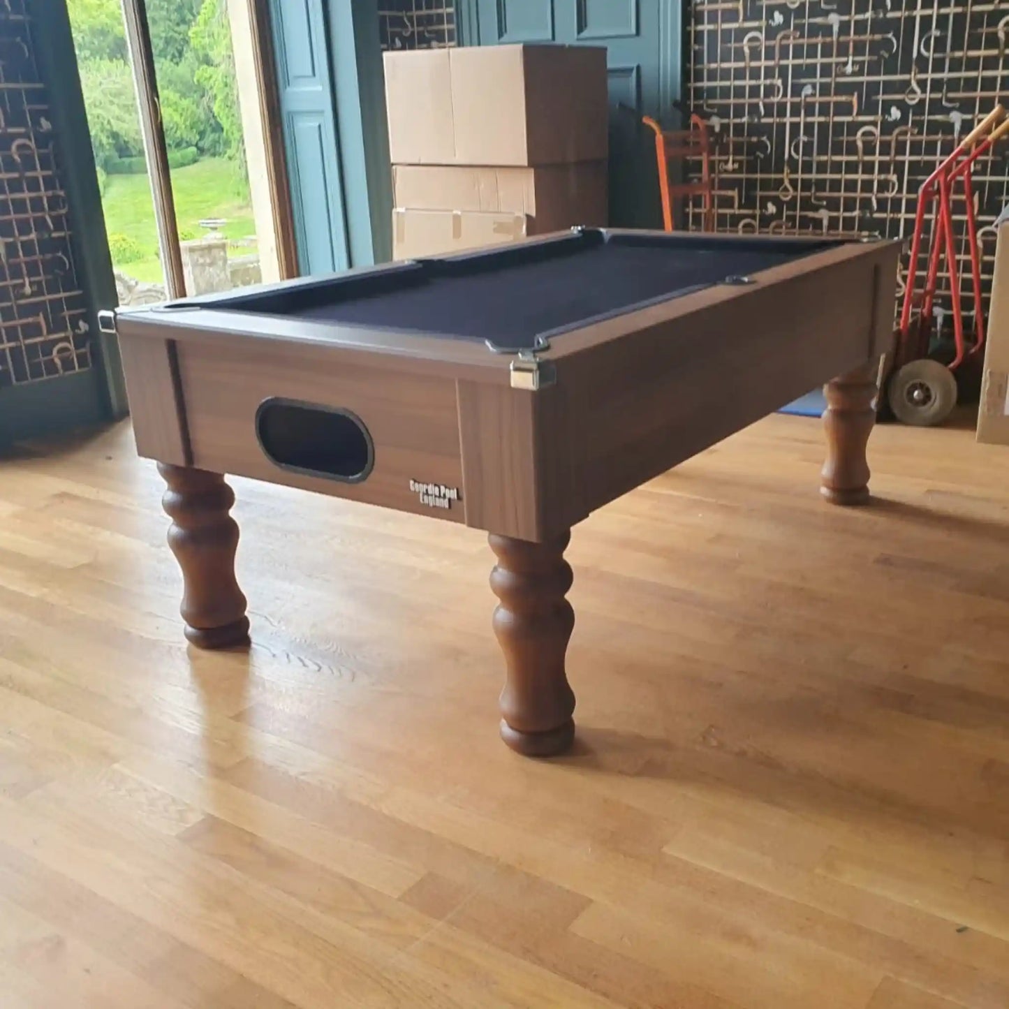 A wooden billiards table with a dark felt playing surface, shown in a room with light wood floors, cardboard boxes, and a tiled wall in the background.