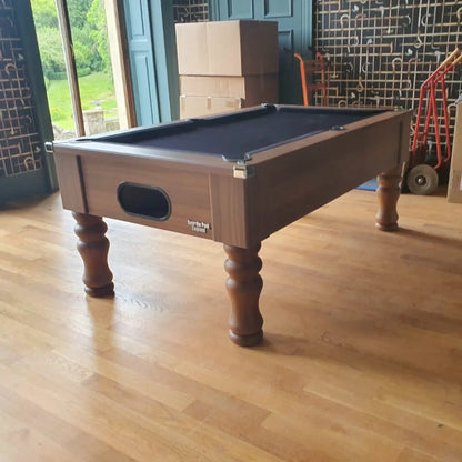 A wooden billiards table with a dark felt playing surface, shown in a room with light wood floors, cardboard boxes, and a tiled wall in the background.