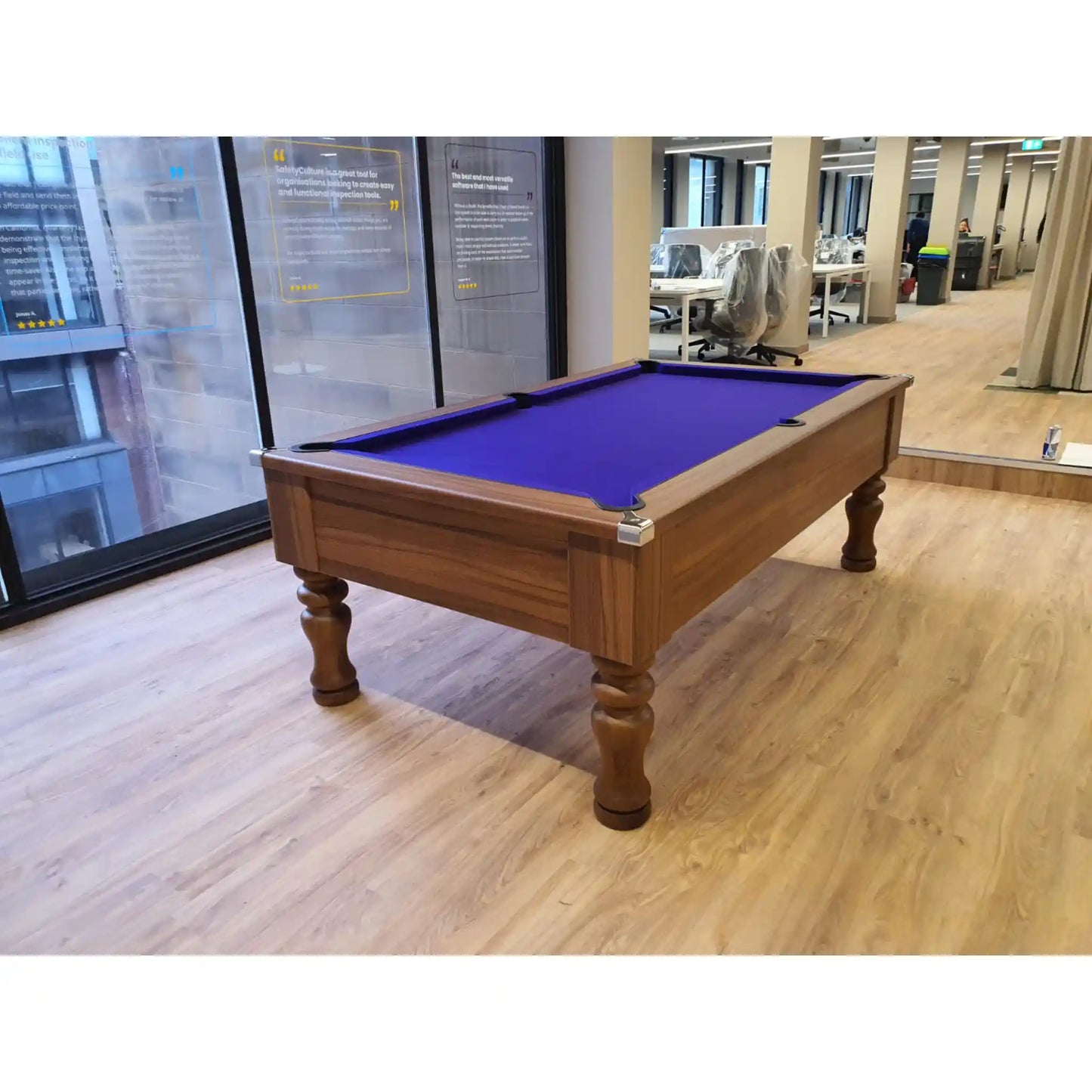 A wooden pool table with a vibrant purple felt playing surface, positioned on a light wood floor inside an office environment.