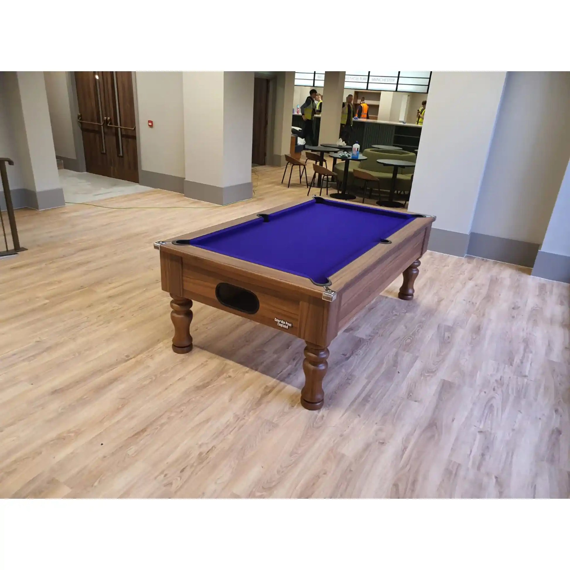 A wooden pool table with a vibrant purple felt playing surface, positioned on a light wood laminate floor in a modern indoor lobby or lounge area.