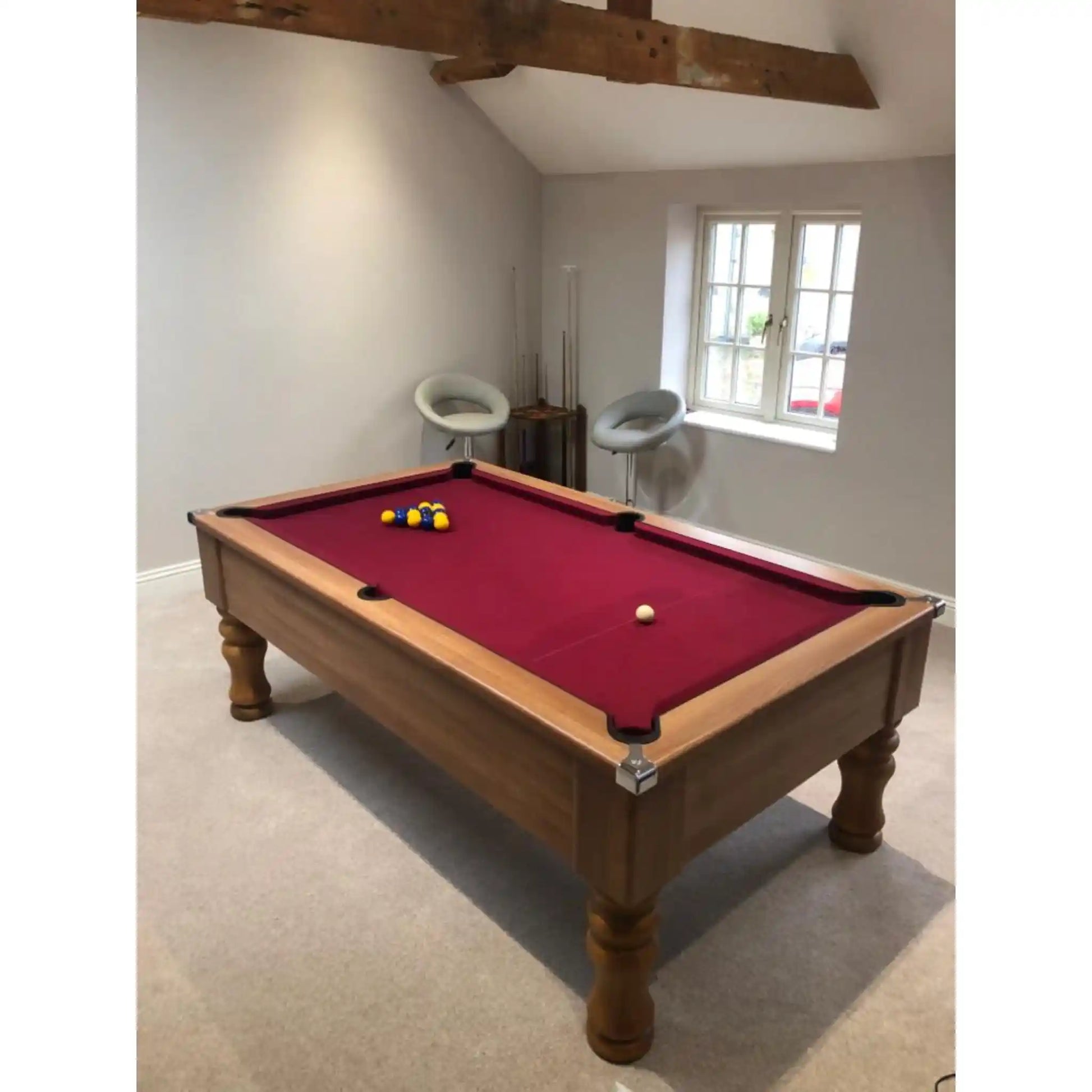 A wooden pool table with a deep red felt surface and billiard balls, set in a room with beige carpet, white walls, and a window.