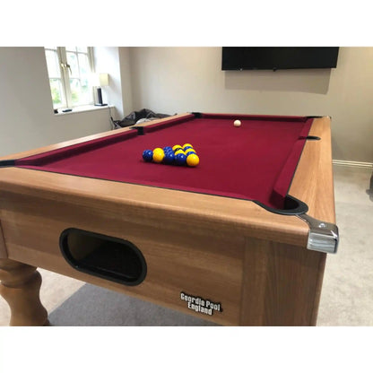 A wooden pool table with a red felt playing surface, blue and yellow billiard balls, and a white cue ball, photographed in a home or studio room with a window, wall-mounted TV, and carpet visible.