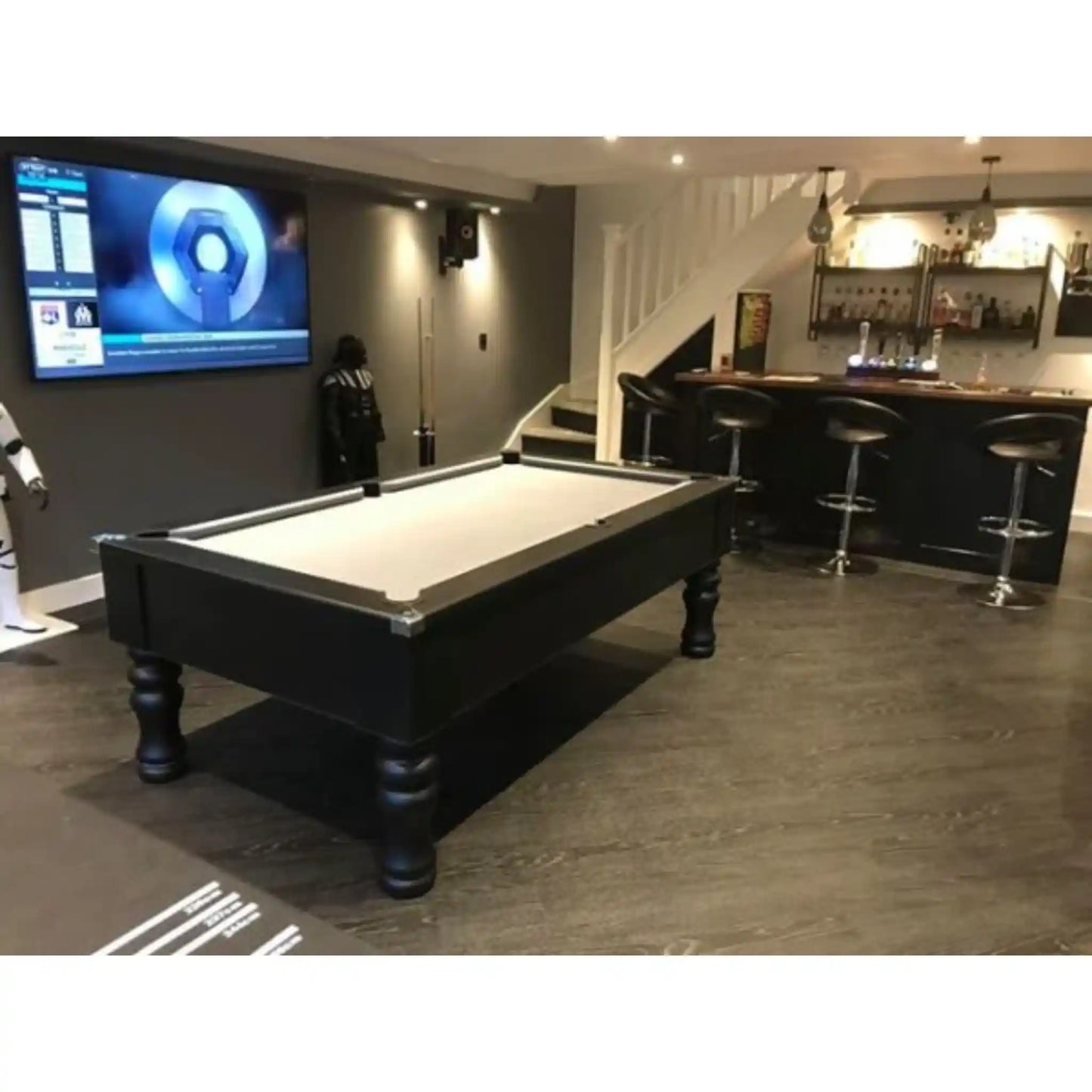 A black pool table with a white felt surface is shown in a modern home bar and recreation room setting with a staircase, bar counter, and bar stools.