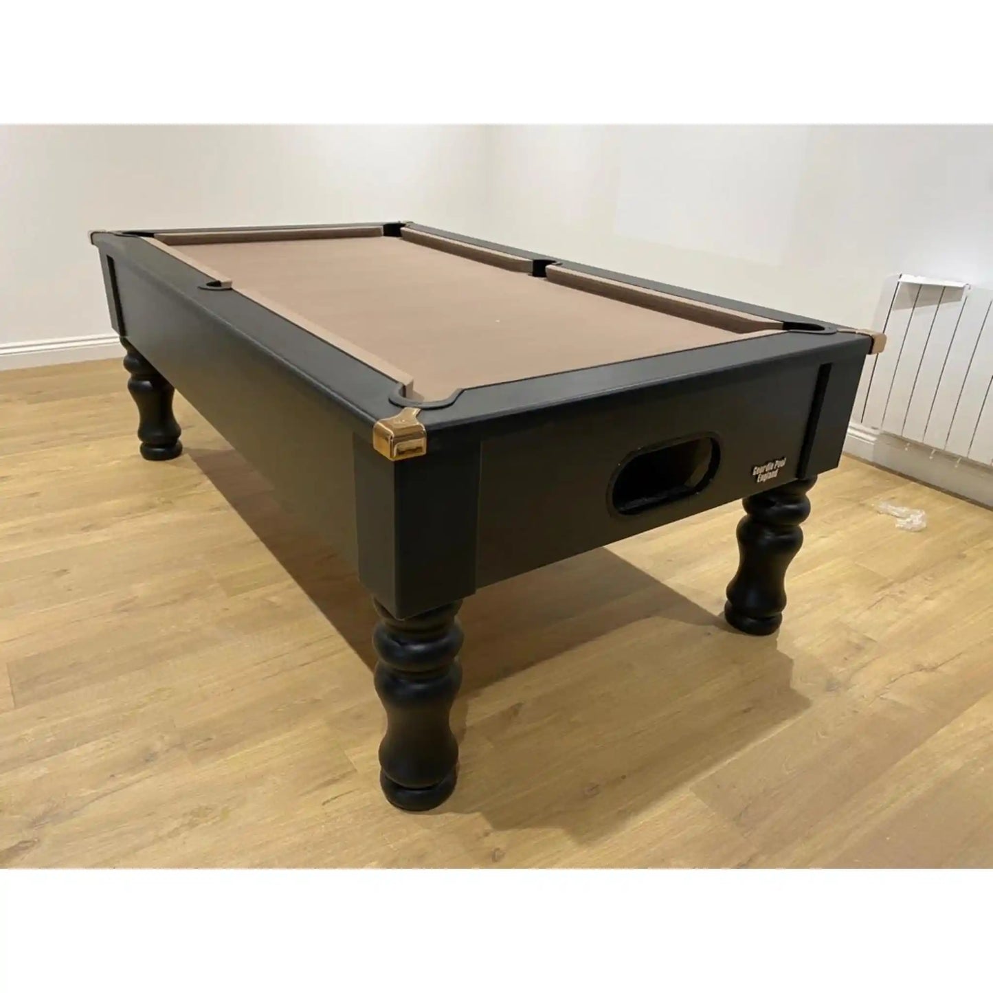 A black wooden billiards table with a tan felt playing surface, shown in a room with light wood floors and off-white walls.