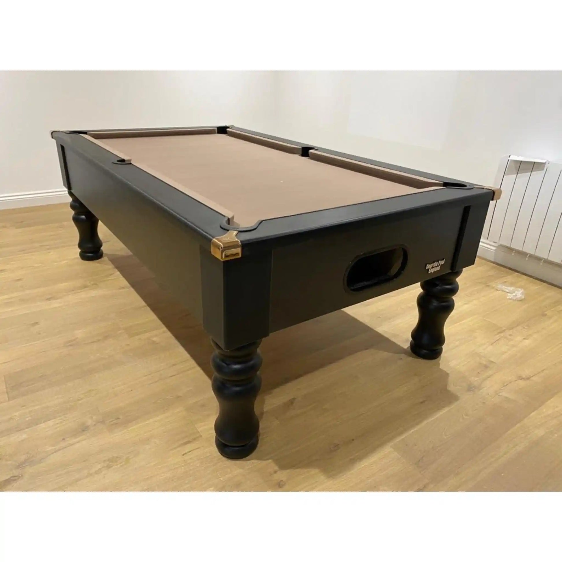 A black wooden billiards table with a tan felt playing surface, shown in a room with light wood floors and off-white walls.