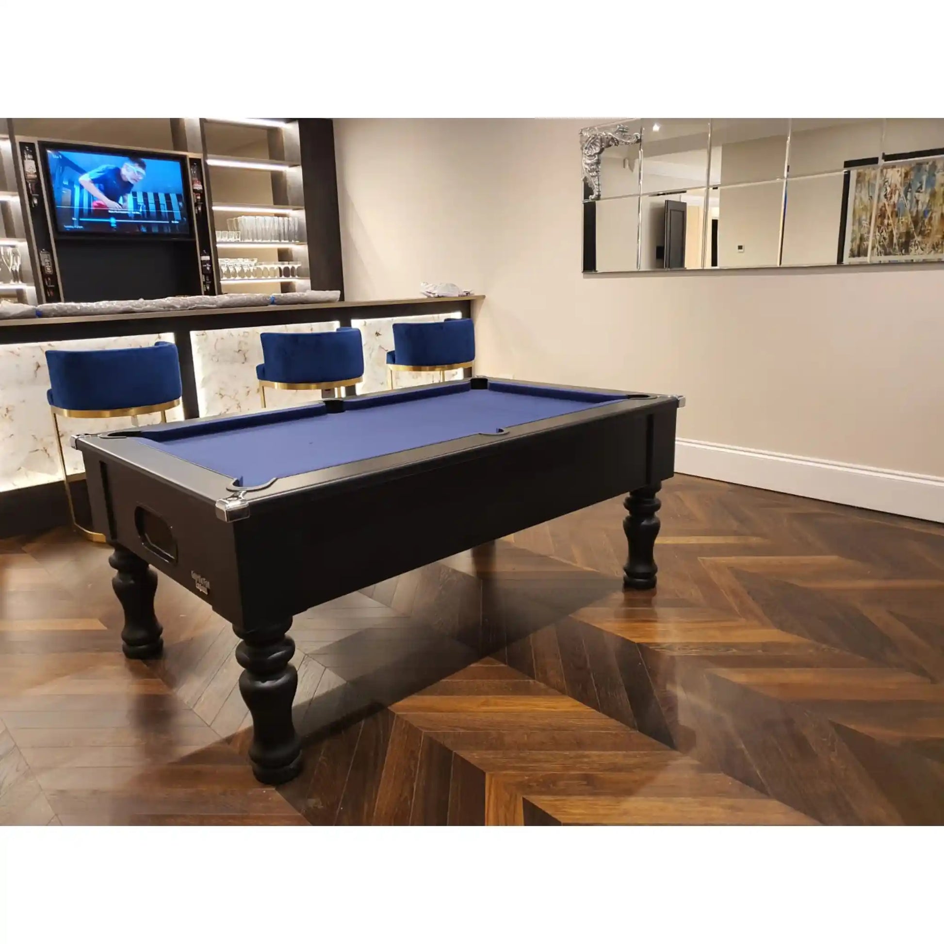A black pool table with a blue felt playing surface, positioned in a modern room with a wood herringbone floor, bar stools, and a television in the background.