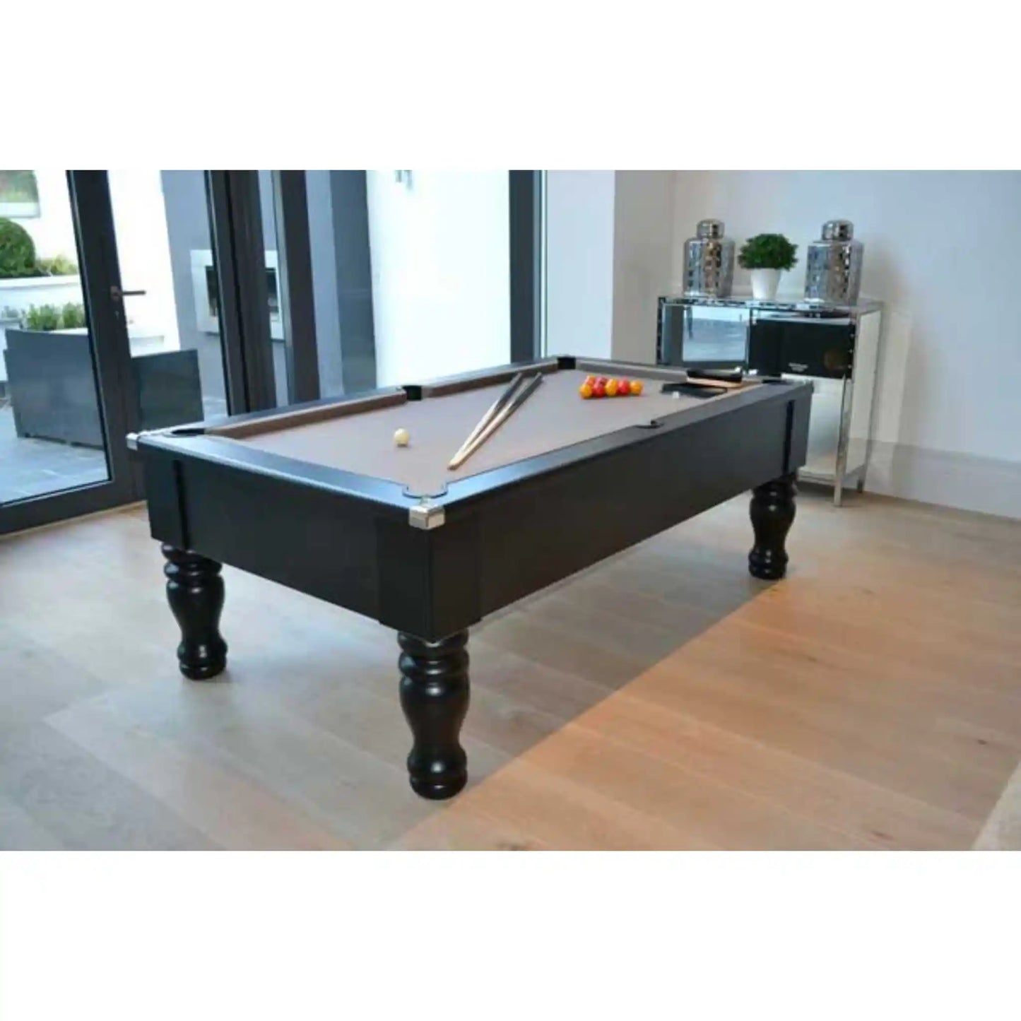 A black pool table with a brown felt playing surface, shown in a modern room with light wood floors and glass doors. The table is set with billiard cues and a rack of colored balls.