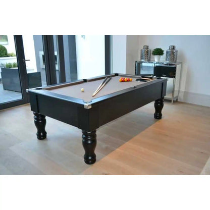 A black pool table with a brown felt playing surface, shown in a modern room with light wood floors and glass doors. The table is set with billiard cues and a rack of colored balls.