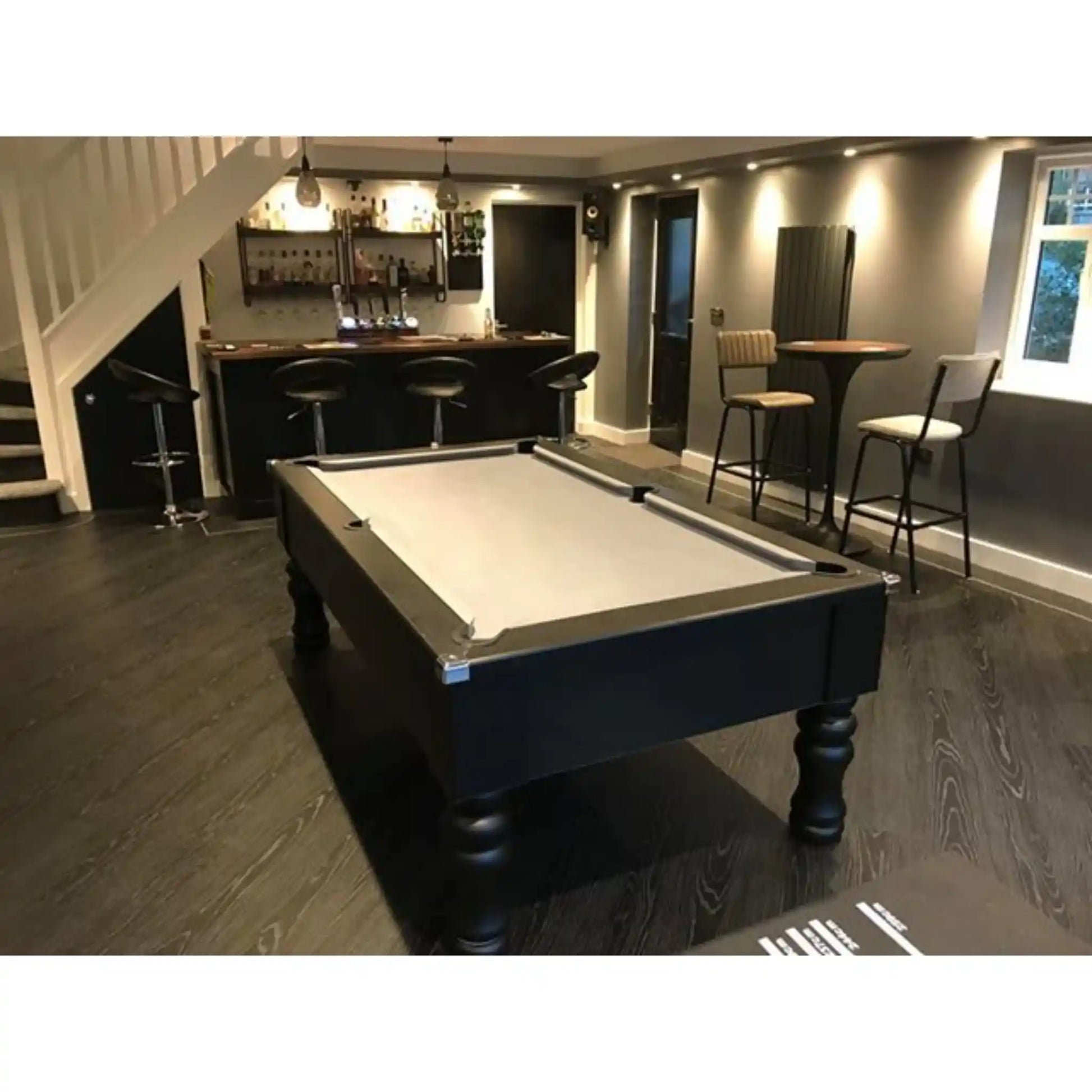 A dark wood pool table with a light-colored felt surface is shown in a home bar and lounge setting with dark wood floors, bar stools, and a staircase in the background.