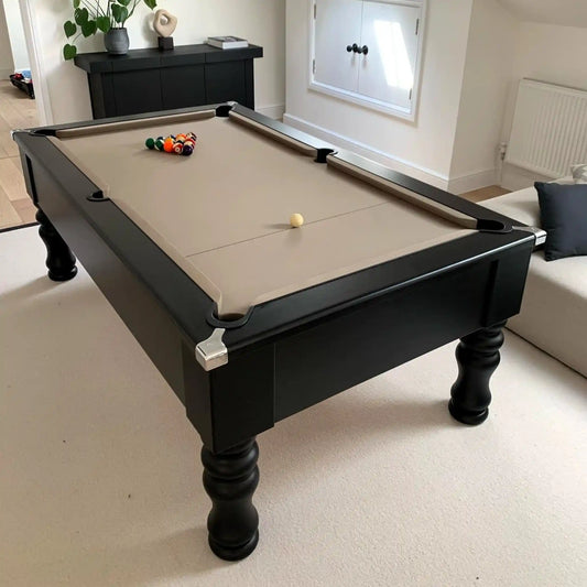 A black wooden pool table with a beige felt playing surface, shown in a bright living room setting. The table is set with a rack of billiard balls and a single cue ball, with a sofa and home decor visible in the background.