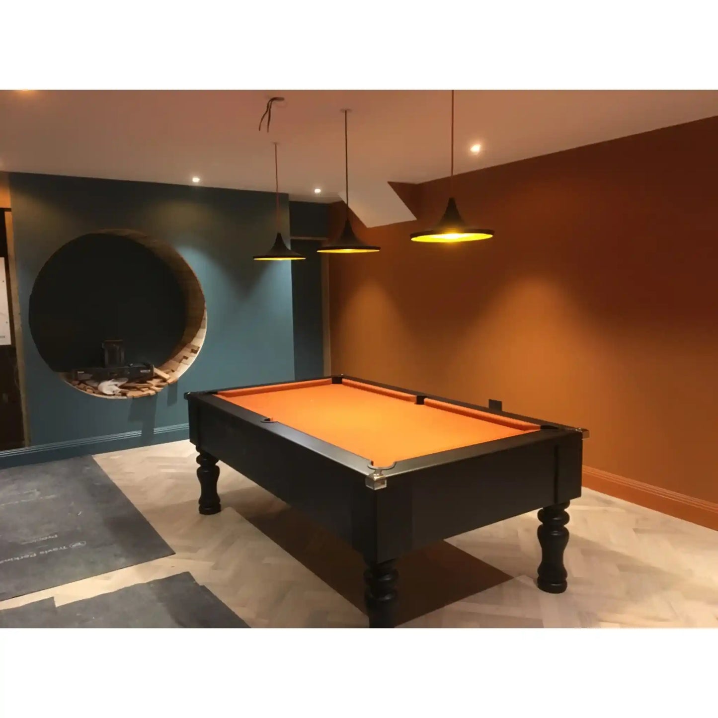 A black pool table with a bright orange felt playing surface, positioned in a modern room with a blue accent wall, a circular window-like feature, and warm brown walls under pendant lights.