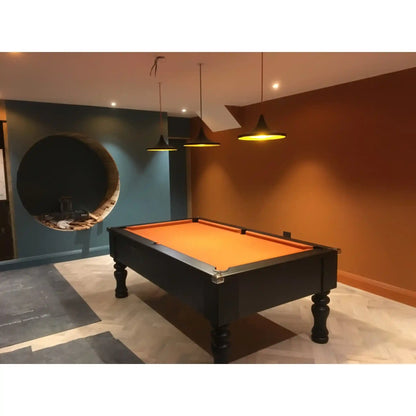 A black pool table with a bright orange felt playing surface, positioned in a modern room with a blue accent wall, a circular window-like feature, and warm brown walls under pendant lights.
