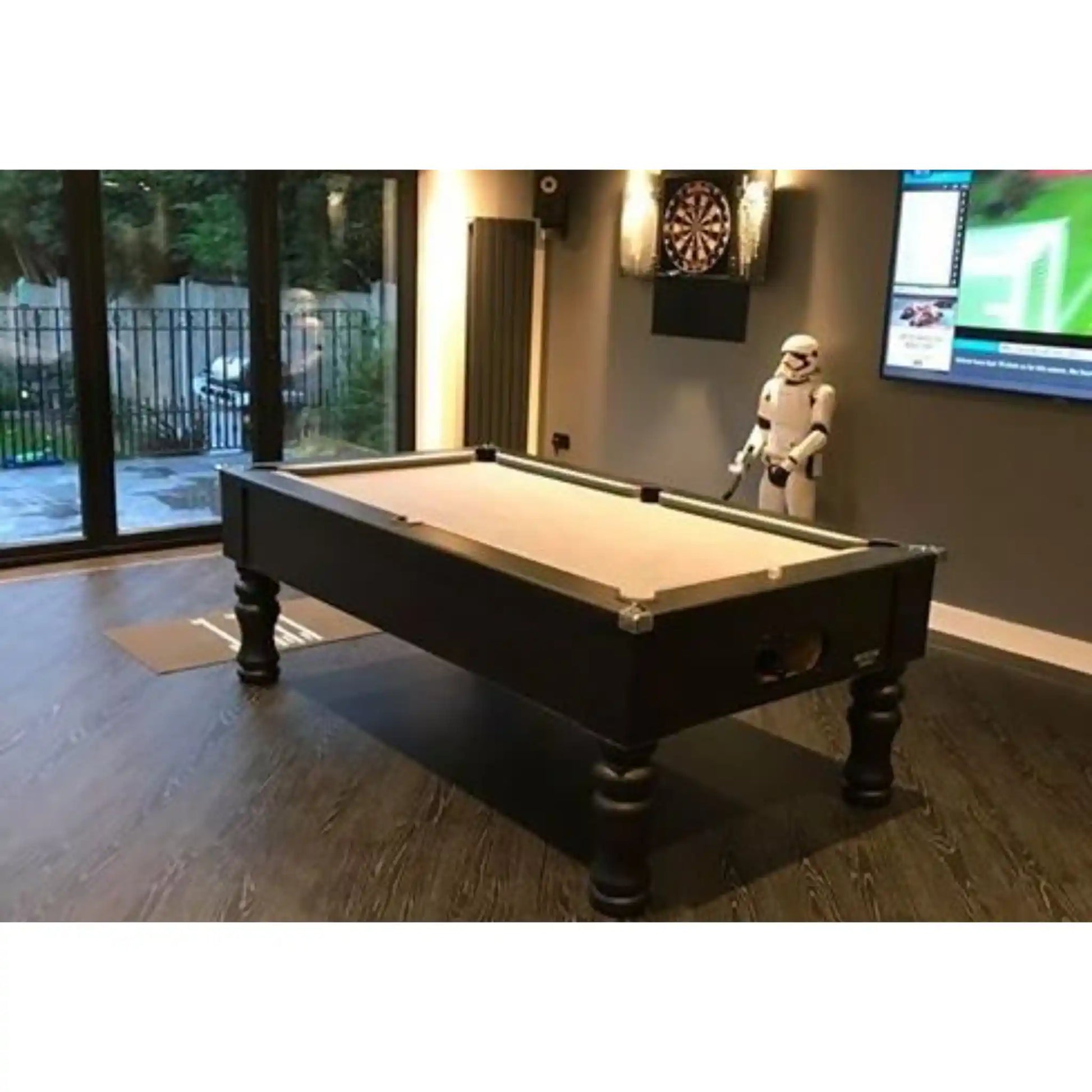 A dark wood pool table with a light beige felt surface in a modern home entertainment room, featuring a dartboard on the wall and a Star Wars Stormtrooper figurine.