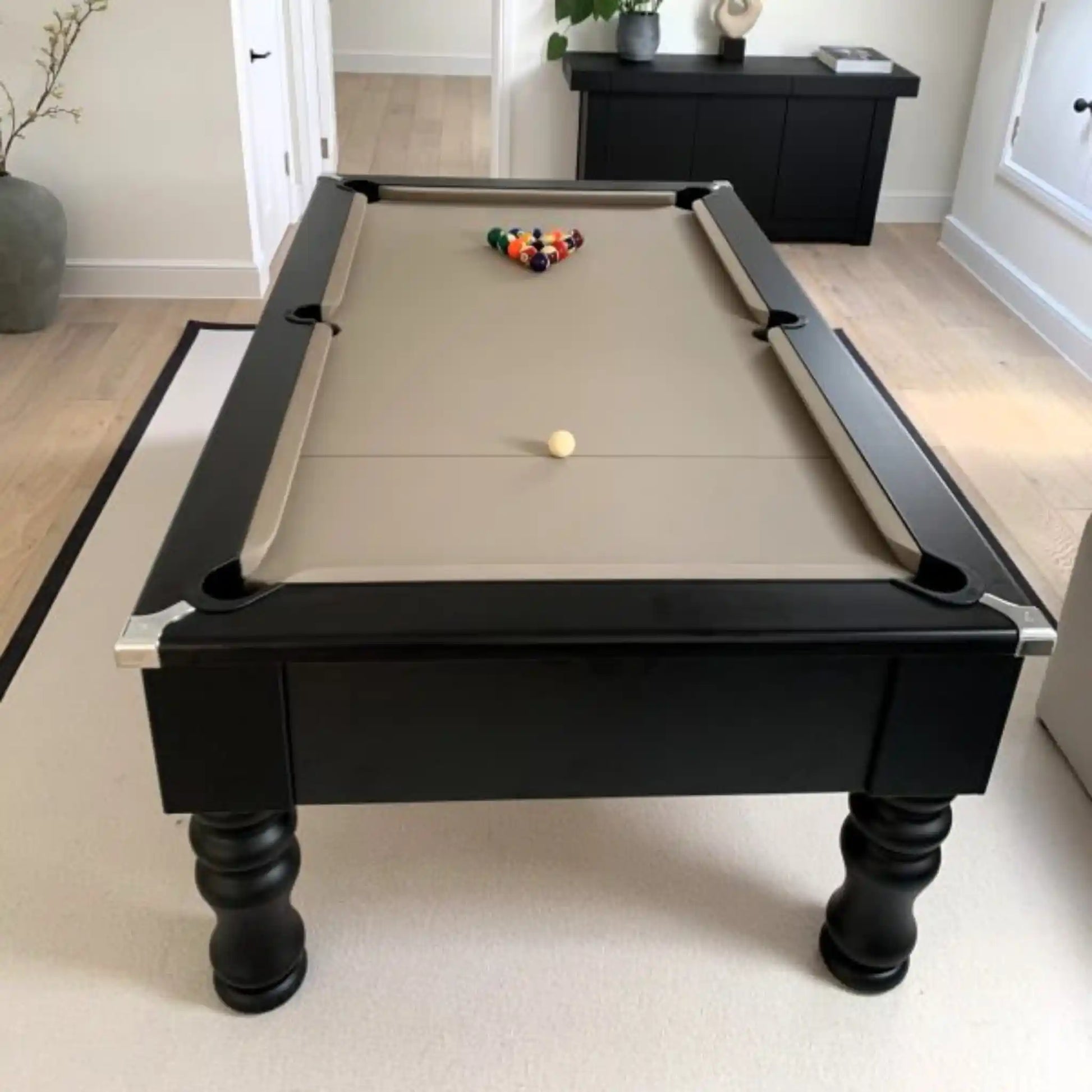 A black pool table with a beige felt playing surface, shown in a bright, modern room with light wood floors and neutral walls. A triangle rack of billiard balls sits on the table, along with a single white cue ball.